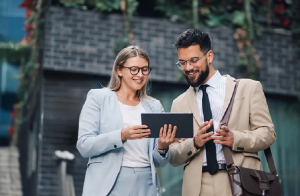 A man and a woman in business suits standing ang and looking at a tablet, smiling