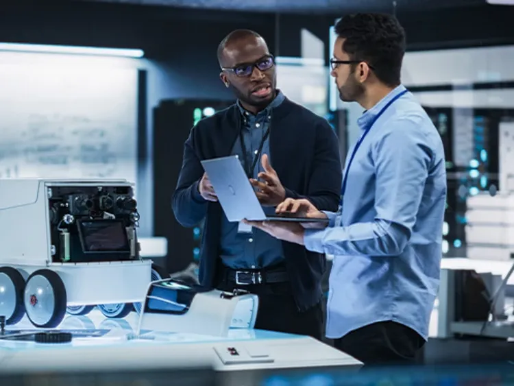 Two male colleagues standing, one talking while the other listens, holding an open laptop