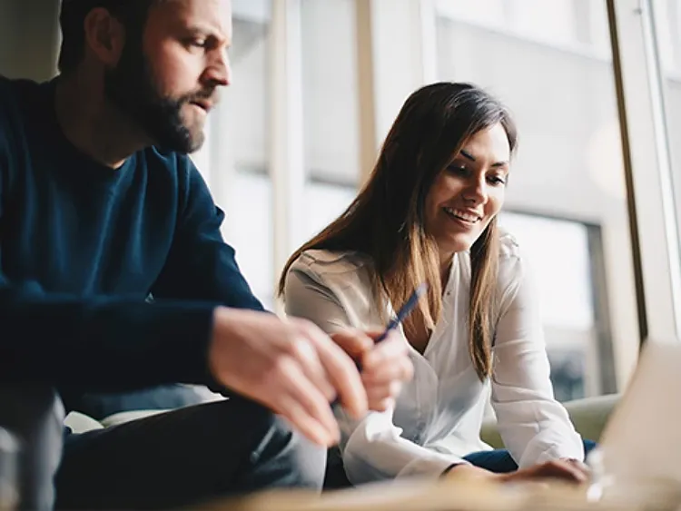 A woman is sitting and working on a laptop, while her partner looks on