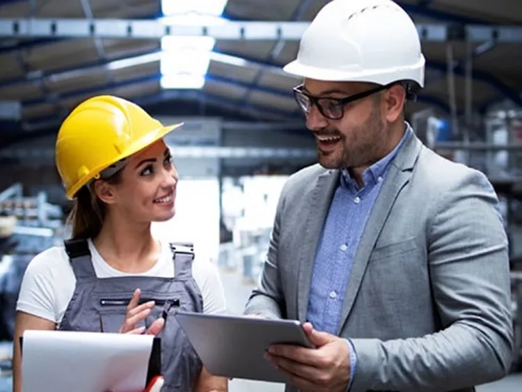 A girl wearing a yellow helmet and gray overalls holding a clipboard listening to a man in a gray suit and white helmet, holding a tablet and talking
