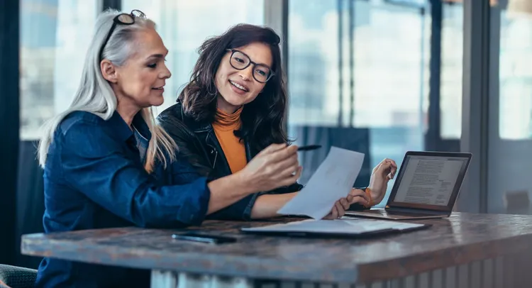 Two women seated at a table, collaborating on a laptop, engaged in discussion