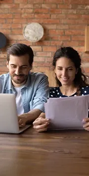 A man and a woman smiling and having a conversation while sitting at a table and looking at documents and a laptop