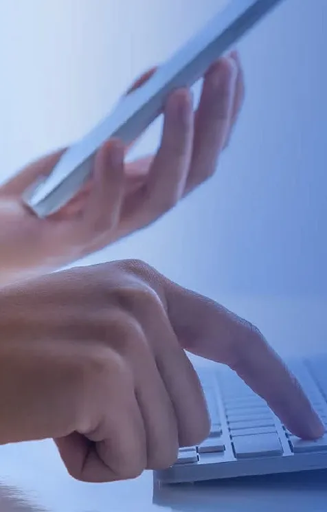 Closeup view of one hand holding cellphone while other hand is typing on keyboard