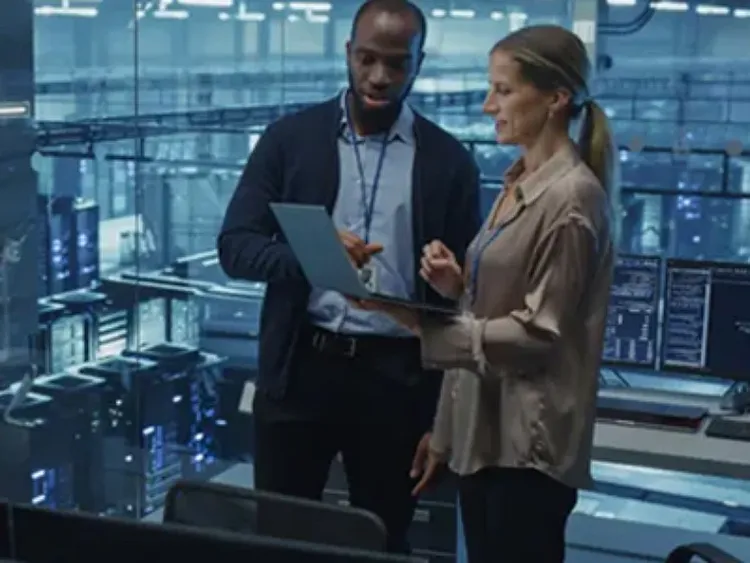 A male holding a laptop along with a female colleague and both are looking into it inside a control room