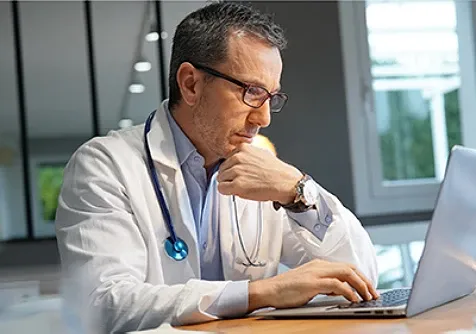 A doctor in a white coat and stethoscope sits at a desk, concentrating on a laptop.