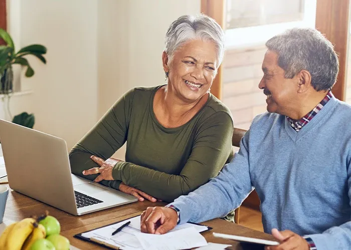 An elderly woman in front of a laptop is smiling at an elderly gentleman sitting next to her