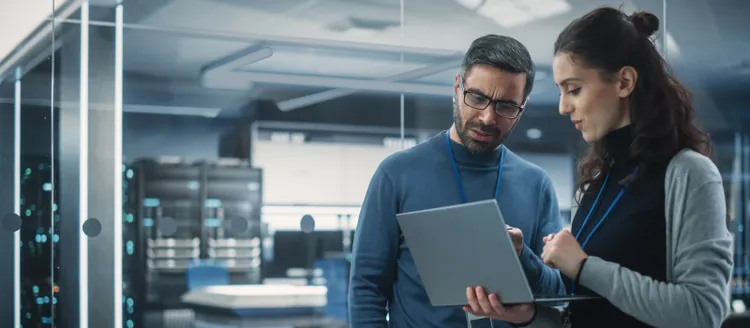 A man and a woman standing and holding a laptop, while discussing something