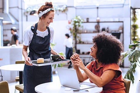 A female server, holding a tray of refreshments, interacting with a client who is seated at a table with an open laptop