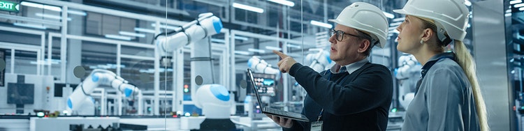 Two individuals in hard hats observe a robot.