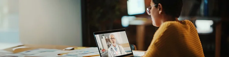 A person wearing glasses and sitting at a table is having a video call with a doctor displayed on a laptop screen.