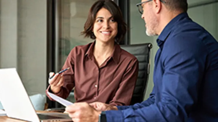 Two colleagues in business attire share a discussion at a desk with a laptop. The woman smiles, creating a positive and collaborative atmosphere.