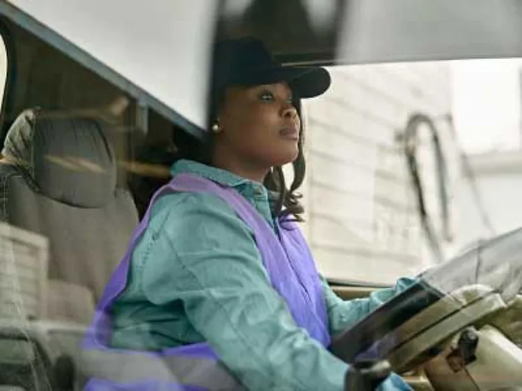 A woman wearing a lavendar vest over a blue shirt, sitting in the driving cabin of a big vehicle