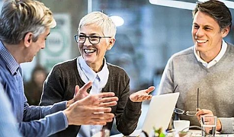 Three people sitting at a table with an open laptop, smiling and discussing something.