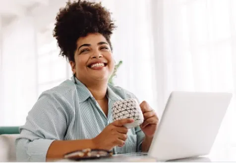 A woman sitting in front of an open laptop, holding a coffee mug and smiling