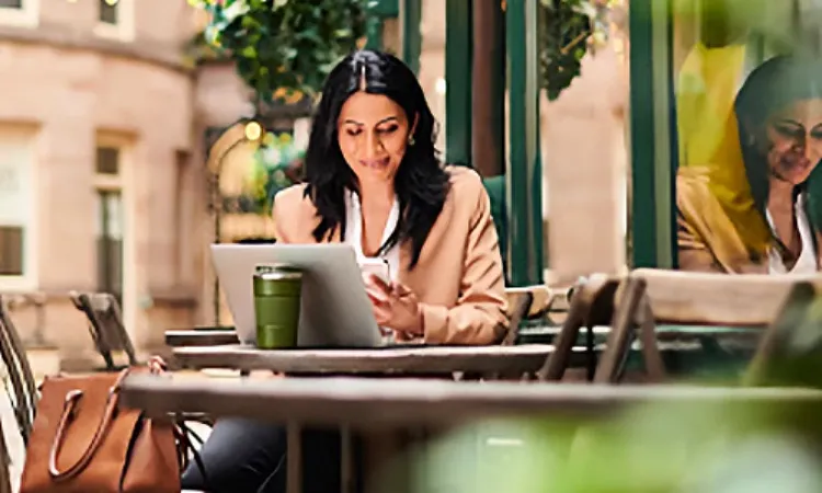 A woman sitting at an outdoor cafeteria with a laptop open before her, smiling and looking at her mobile phone
