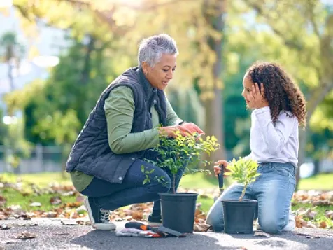 A woman sitting on her haunches with two potted plants explaining something to a little girl kneeling and listening intently