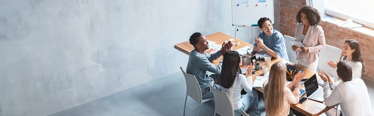 A professional woman is standing with a notepad in her hand while her colleagues clap for her sitting around an office table