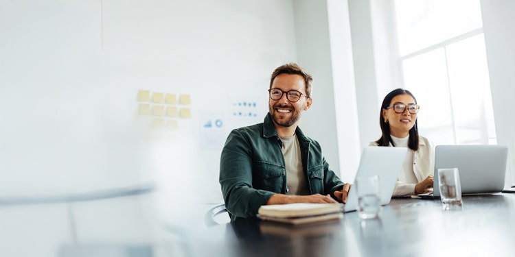 Two people sitting at a table with open laptops in front of them, while they are looking at something outside the frame and smiling