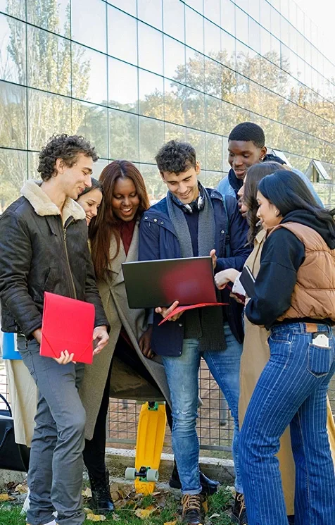 A group of young adults huddled around a young man holding an open laptop and looking at it together