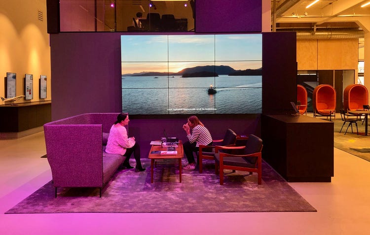 Two women sitting at a lounge-like office space that has a big digital screen on the wall